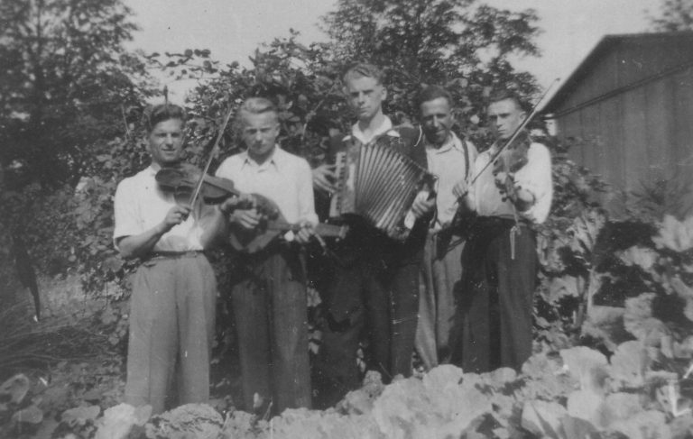 Aufnahme von ca. 1943 vor der Baracke in der Uetersener Straße in Tornesch. Fünf Personen sind abgebildet, zwei davon mit Geigen, ein Akkordeon und ein Mandolinenspieler. Von links: zwei Ukrainer Leotz und Joseph, dann der Pole Henryk Bejerski, dann die beiden Ukrainer Dimit Noworad und Kasimirsch. Sie machten privat sonntags Musik. Foto von Hans-Joachim Wohlenberg.